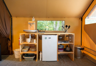 Keukengedeelte in een safaritent met koelkast, wasbak, servies, pannen en keukengerei op planken.