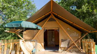 Tenda safari con terrazza in legno, ombrellone verde e sedia da campeggio circondata da alberi verdi.