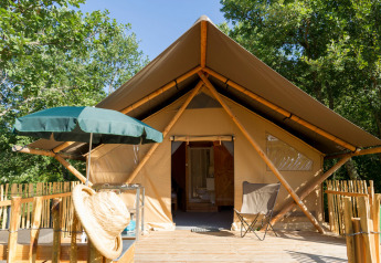 Tenda safari con terrazza in legno, ombrellone verde e sedia da campeggio circondata da alberi verdi.