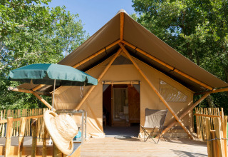 Safarizelt mit Holzterrasse, grünem Sonnenschirm und Campingstuhl inmitten grüner Bäume bei Sonne.