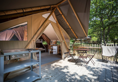 Safari-Zelt mit Holzterrasse, Outdoor-Möbeln, Gaskocher und Blick auf grüne Bäume im Freien.