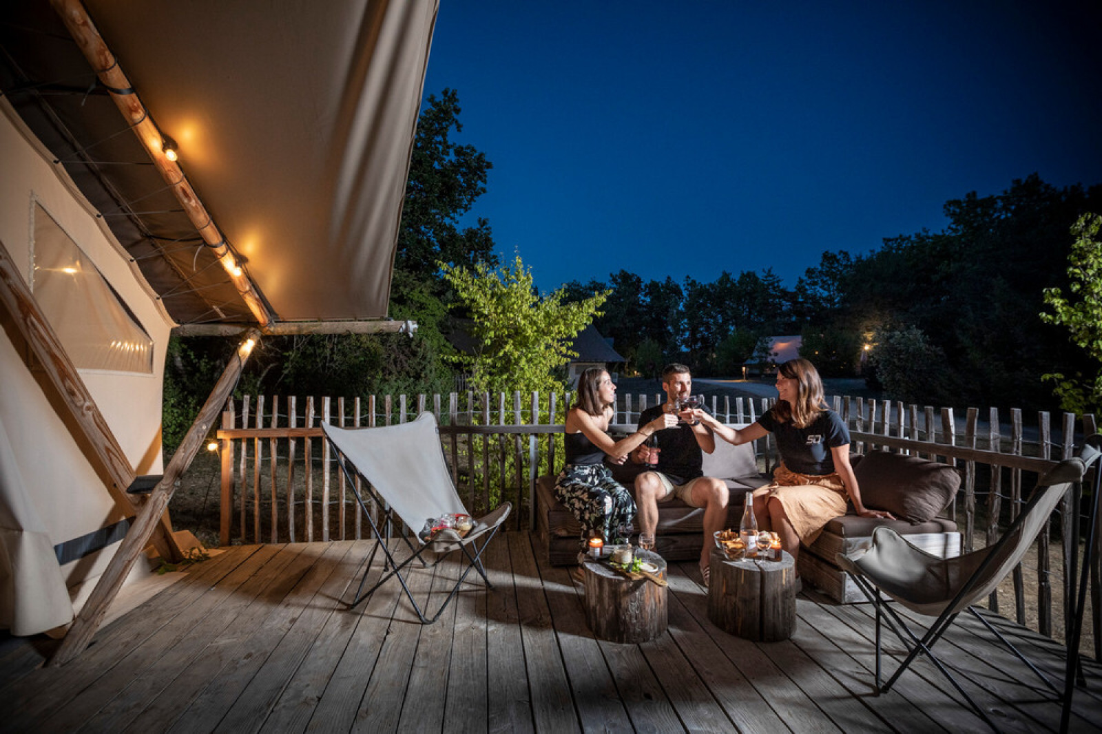 Drie mensen genieten van drankjes op het verlichte terras van een safaritent op een zomerse avond.