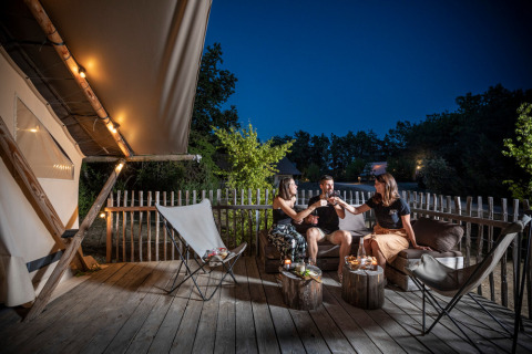 Tre personer nyder drinks på terrassen foran et oplyst safaritelt ved aften under åben himmel.