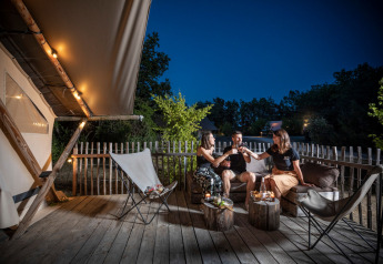 Tre persone brindano con drink sulla terrazza illuminata di una tenda safari durante una sera estiva.