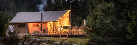 Tienda safari con luces y chimenea en un bosque al anochecer, rodeada de árboles y rocas grandes.