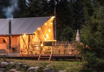 Safari tent with lights and chimney in a forest setting at dusk, surrounded by trees and large rocks.