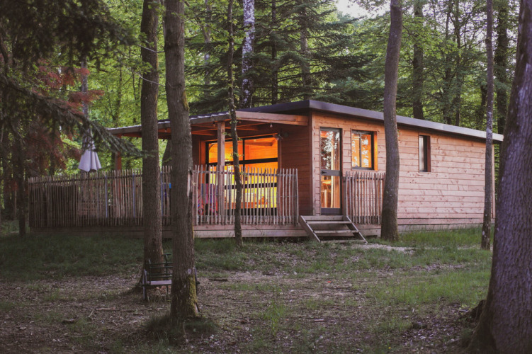 Cabaña de madera Chalet Toronto con luces cálidas en el bosque de Village Huttopia Pays de Condrieu, Francia, al anochecer.