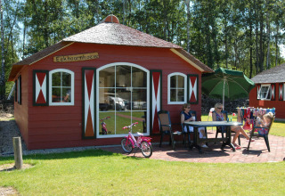 Red chalet marked Eekhoornhoeve with three people relaxing outside at Veluwecamping De Pampel, Gelderland.