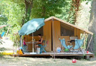 Gezin geniet van camping voor een grote tent met tuinmeubilair onder bomen in een groene omgeving.