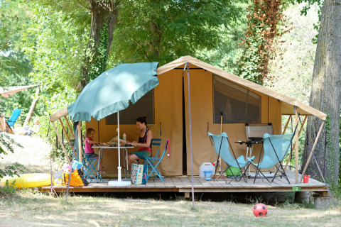 Gezin geniet van camping voor een grote tent met tuinmeubilair onder bomen in een groene omgeving.