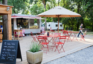 Buitencafé met rode tafels en stoelen, parasol en foodtruck bij Huttopia Millau, Occitanië, Frankrijk.
