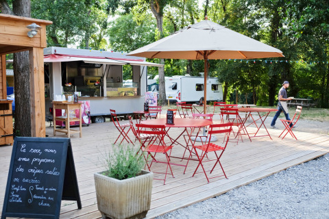 Outdoor café with red tables and chairs, umbrella, food truck and greenery at Huttopia Millau holiday park.