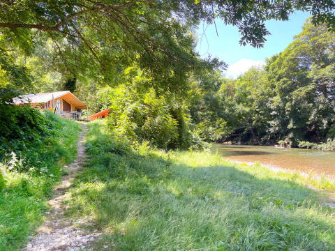 Ein Pfad führt zu einem Zeltlager am Fluss, umgeben von Bäumen im Huttopia Millau, Occitanie, Frankreich.