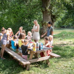 Niños y adultos reunidos alrededor de una mesa de picnic en el parque verde de Huttopia Millau, Occitania, Francia.