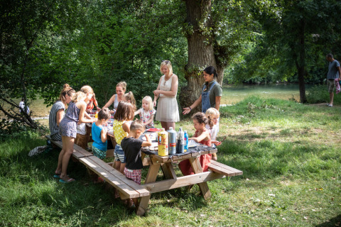 Niños y adultos reunidos alrededor de una mesa de picnic en el parque verde de Huttopia Millau, Occitania, Francia.
