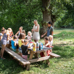 Niños y adultos reunidos alrededor de una mesa de picnic en el parque verde de Huttopia Millau, Occitania, Francia.
