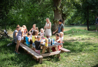 Bambini e adulti riuniti attorno a un tavolo da picnic nel parco verde di Huttopia Millau, Occitania, Francia.