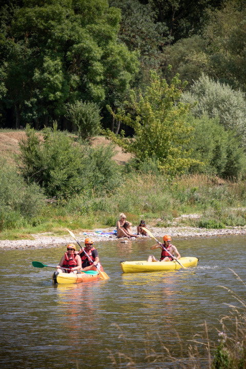 To personer sejler i kajak på en flod ved Huttopia Millau, mens to andre slapper af på bredden i Occitanie.