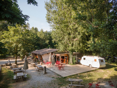 Área de cafetería al aire libre con mesas de picnic y caravana entre árboles en Huttopia Millau, Occitanie, Francia.