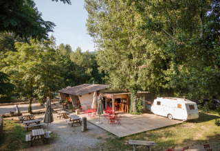Udendørs caféområde med borde, stole og campingvogn omgivet af træer i Huttopia Millau, Occitanie, Frankrig.