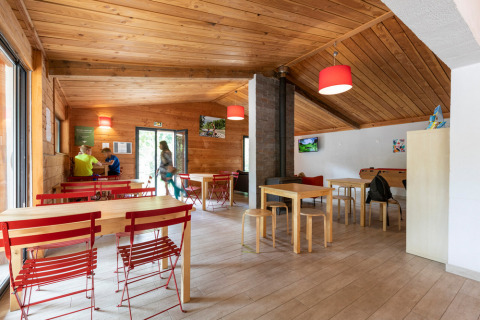 Area comune interna con arredi in legno, sedie rosse e ospiti in un villaggio vacanze in Occitania, Francia.