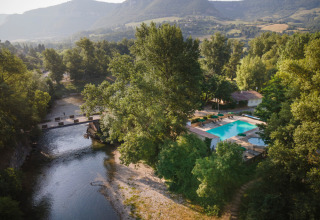 Aerial view of Huttopia Millau holiday park in Occitanie, France, featuring a pool, river, and lush mountains.