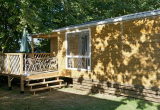 Mobilhjem Cottage ved Huttopia Millau i Frankrig, med terrasse, stole og grøn parasol under træer.