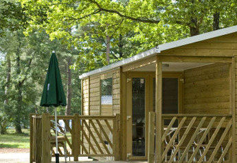 Mobilhytte Cottage ved Huttopia Millau i Frankrig, omgivet af træer og med en træveranda og parasol.