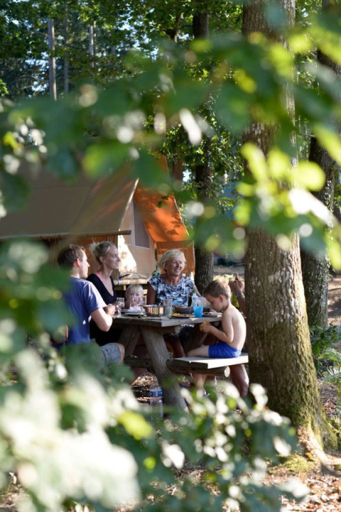 Family enjoys a meal at a picnic table in the woods at Village Huttopia Senonches, Centre-Val de Loire, France.
