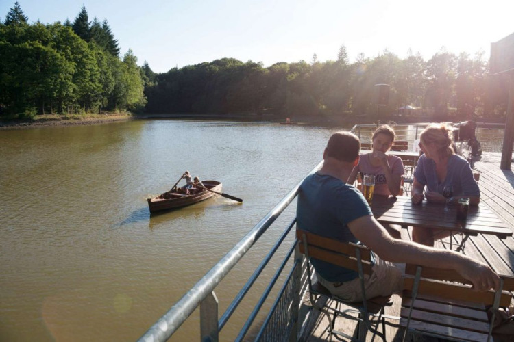 People relaxing with drinks on a lakeside terrace at Village Huttopia Senonches, Centre-Val de Loire, France.