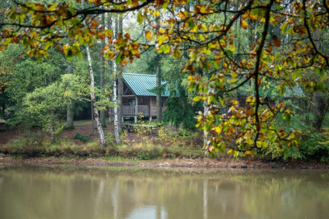 Cabaña de madera con techo verde junto a un lago en Village Huttopia Senonches, rodeada de bosque frondoso.