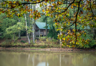 Houten hut met groen dak aan een meer in Village Huttopia Senonches, omgeven door dicht bosrijk gebied.
