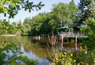 Vue sur le lac au Village Huttopia Senonches, avec une terrasse, une barque et de grands arbres verts.