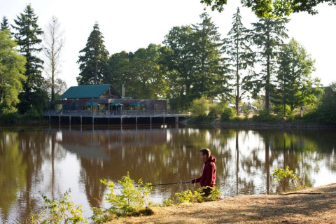 Hombre pescando junto a un lago con cabaña y árboles al fondo en Village Huttopia Senonches, Francia.