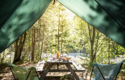 Vista desde el interior de una tienda tipi llamada Bonaventure hacia una mesa de picnic y bosque soleado.