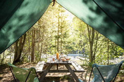 Vista desde el interior de una tienda tipi llamada Bonaventure hacia una mesa de picnic y bosque soleado.