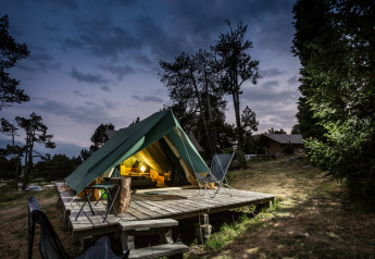 Das beleuchtete Teepee-Zelt Bonaventure steht abends auf einer Holzplattform im Wald mit Stühlen davor.