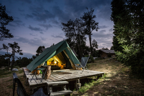 The Bonaventure teepee tent lit up at night on a wooden platform in the woods, with chairs outside.