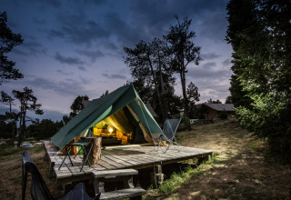 La tienda tipi Bonaventure iluminada por la noche sobre una plataforma de madera en el bosque, con sillas afuera.