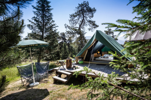 Tienda tipi Bonaventure sobre plataforma de madera en el bosque, con sillas, sombrilla y montañas.