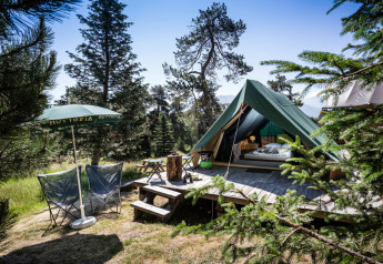 Bonaventure-Tipizelt auf Holzdeck im Wald, mit Stühlen, Sonnenschirm und Bergen im Hintergrund.
