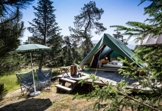 Tienda tipi Bonaventure sobre plataforma de madera en el bosque, con sillas, sombrilla y montañas.