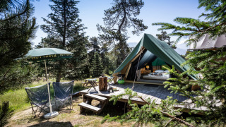 Bonaventure-Tipizelt auf Holzdeck im Wald, mit Stühlen, Sonnenschirm und Bergen im Hintergrund.