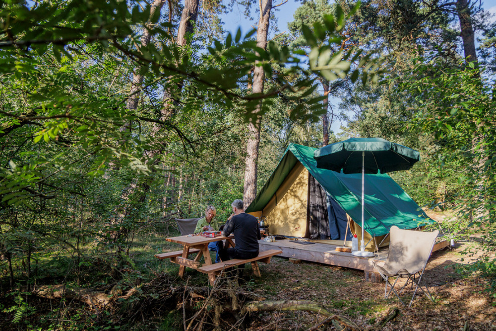Twee mensen zitten aan een picknicktafel bij een Bonaventure tipi tent in een groene bosrijke omgeving.