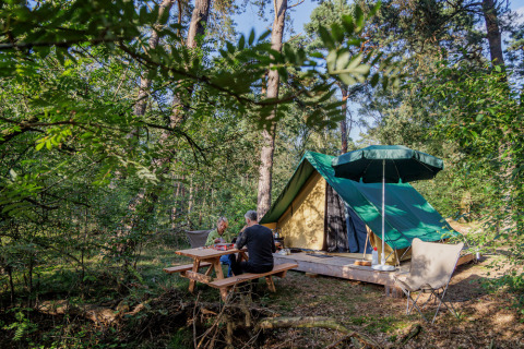 Dos personas sentadas en una mesa de picnic frente a una tienda tipi Bonaventure en un bosque verde.