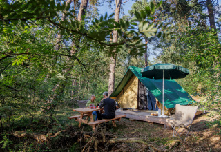 To personer nyder et måltid ved et picnicbord foran en Bonaventure tipitelte i en grøn skov.