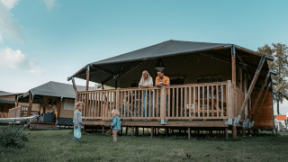 Familia relajándose en la terraza de una cabaña mientras dos niños juegan en el césped frente al lodge.