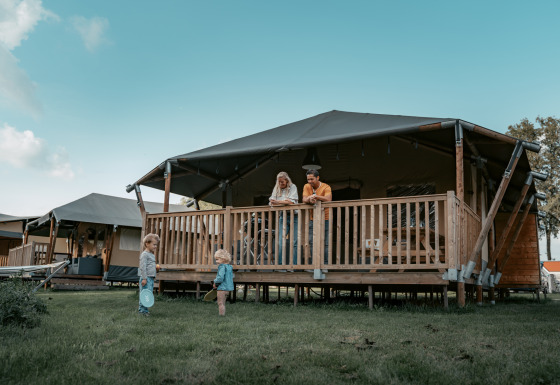 Familia relajándose en la terraza de una cabaña mientras dos niños juegan en el césped frente al lodge.