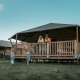 Family relaxing on the porch of a wooden cottage lodge, while two children play on the grass in front.