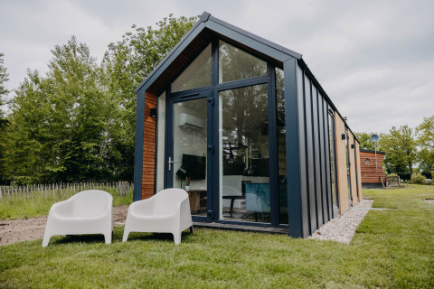 Modernes Tiny House Oslo Suite mit großen Fenstern, Rasenfläche und zwei weißen Stühlen davor.